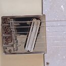 Detailní pohled na chromovaný kobylku a snímač elektrické kytary na krémovém povrchu s bílým perleťovým pickguardem. Kobylka je kovová, s viditelnými…