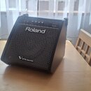 Černý kombozesilovač značky Roland, model PM-100. Na přední mřížce je nápis Roland a v dolní části označení V-Drums. Horní panel obsahuje ovládací pr…