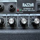 Detailní pohled na ovládací panel kytarového komba s černým texturovaným povrchem. Na panelu je nápis RAZZOR STREET-5 GUITAR AMPLIFIER, vypínač POWER…