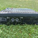 Černý mixážní pult s řadou stříbrných a barevných ovladačů, umístěný na trávníku. Na zadním panelu je vidět nápis YAMAHA POWERED MIXER a označení mod…