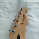 Detail hlavy elektrické kytary z přírodního světlého dřeva. Na povrchu je hnědý nápis Fender a svislý text TELECASTER. Jsou vidět šest chromovaných l…