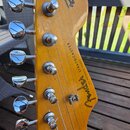 Fotografie zachycuje hlavu elektrické kytary z vybarveného dřeva s šestnácti chromovanými ladičkami. Na povrchu je černý nápis Fender a STRATOCASTER,…