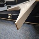 Detailní pohled na bílé tělo elektrokitary v černém kufru. Na spodním špičatém okraji těla je viditelné výrazné opotřebení laku a odloupaný povrch, p…