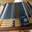 Míčovací pult s černým povrchem a dřevěným rámem. Na přístroji je viditelný nápis Model 24 a logo TASCAM. Obsahuje řadu bílých posuvníků, barevné oto…