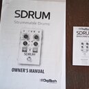 Fotografie zobrazuje dva papírové dokumenty na hnědém dřevěném podkladu. Vlevo je větší příručka s nápisy SDRUM, Strummable Drums™, OWNER'S MANUAL a…