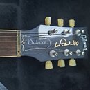 Detail hlavy elektrokitary v černé barvě s bílými nápisy. Na hlavě jsou doslovně uvedeny názvy Deluxe, Leo Paul 100 a Gibson. Nástroj má šest ladicíc…