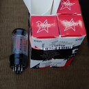 Na fotografii je elektronická lampa s černým plastovým základním soketem a červenými nápisy na skle. Vedle lampy jsou tři červené kartonové krabičky…