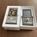 Dvě bílé krabice, v nichž je umístěn stroboskopický ladicí přístroj v šedé barvě. Na zařízení je uveden nápis STROBOSTOMPHD peterson STROBE TUNERS a…