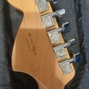 Záběr na zadní stranu hlavy kytary z přírodního světlého dřeva. Na dřevě je vyryto „SERIAL NUMBER US11134258“ a pod tím nápis „MADE IN USA“. Hlava je…