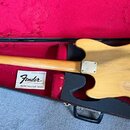 Elektrická basová kytara s přírodním dřevěným povrchem v černém kufru s červenou výstlkou. Na hmatníku je vidět nápis Fender MUSICMASTER BASS. Tělo i…