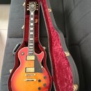 Elektrická kytara typu Les Paul s červeno-žlutým sunburst povrchem v černém tvrdém kufru s červeným plyšovým vnitřkem. Na hlavě hmatníku je nápis Gib…