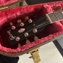 Detail hlavy elektrické kytary v hnědém kufru s červeným plyšovým vnitřkem. Na černé hlavě je bílý nápis Gibson a logo ve tvaru šipky. Jsou vidět kov…