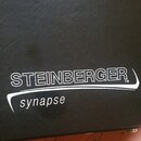 Detailní záběr na černý povrch z texturovaného materiálu připomínajícího kůži. Na povrchu je bílý nápis STEINBERGER TM a pod ním v oblouku slovo syna…
