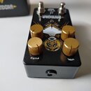 Černý efekt pedál s nápisem PANDORA a grafickým motivem v podobě stylizované masky. Na horní straně jsou čtyři zlaté kovové oválné knofíky a dva kovo…