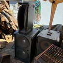 Na fotografii jsou tři černé reproduktorové bedny a míchací pult. Horní reproduktor je umístěn svisle, spodní dvě bedny vedle sebe; všechny mají čern…