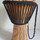 Djembe s tělem z vyřezávaného světlého dřeva a napnutou kůží v horní části. Nástroj je opleten černými provázky, které drží membránu. Spodní část kor…