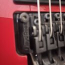 Detailní pohled na černý kovový kobylku elektrické kytary s červeným povrchem těla. Na boku kobylky je vyryt nápis Floyd Rose. Jsou vidět tři kovové…