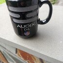 Černý keramický hrnek s úchytem. Na povrchu je stříbrný grafický prvek připomínající mřížku mikrofonu a bílý nápis AUDIX. D6 spolu s malou vlajkou US…