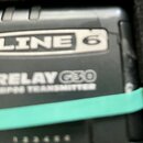 Detailní záběr na černý plastový bezdrátový vysílač. Na zařízení je bílý nápis LINE 6 v obdélníkovém rámečku a pod ním text RELAY G30 a TP-06 TRANSMI…
