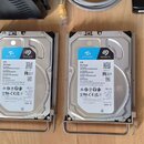 Čtyři stříbrné pevné disky v šedých kovových držácích. Každý disk má bílý štítek s modrým logem a nápisy SKYHAWK, SEAGATE, 6TB SkyHawk. Na štítcích j…