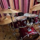 Akustická bicí souprava v tmavě červené barvě s kovovým hardwarem. Na jednom z činelů je nápis Paiste, na korusech a basovém bubnu jsou viditelná log…