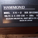 Detailní záběr na černý štítek s technickými údaji na černém povrchu přístroje. Na štítku je uveden název HAMMOND, MODEL XK-2 a SER. 00122995. Dále j…