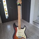 Elektrická kytara typu Stratocaster s javorovým hnedým krkem a tělem v sunburst provedení. Na hlavě krku je svisle napsaný nápis Squier. Kytara má bí…