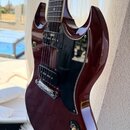Tmavě červená elektrická kytara s černým pickguardem a hnedým hmatníkem. Na těle jsou vidět dva snímače typu humbucker, stříbrný kobylník a čtyři čer…