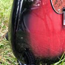 Detailní záběr na červeně a černě lakované tělo elektrické kytary s glossy povrchem. Na laku jsou vidět četné drobné škrábance a stopy opotřebení. V…