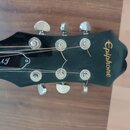 Černý hlavník kytary s nápisem Epiphone v žluté barvě. Na levé straně je ručně napsaný bílý podpis AJ. Jsou vidět šest kovových ladiček a struny, z n…