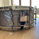 Šedý snare drum s kovovým korpusem s kovaným povrchem a chromovanými obručemi a lugy. Na chromovaném držáku je vyryto logo znacky DW. Nástroj vykazuj…