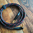 Černý opletený kabel s červenými detaily, srolovaný do kruhu na dřevěném povrchu. Kabel má na koncích zlaté 6,3mm jack konektory s černými plastovými…