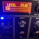Detailní pohled na ovládací panel černého elektronického zařízení. V horní části je červeně podsvícený displej s nápisy LEVEL 00dB, IN1 a IN2. Panel…