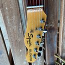 Fotografie zachycuje hlavu elektrické kytary z přírodního dřeva s nápisy Squier, TELECASTER a menším nápisem by Fender. Na hlavě jsou umístěny šest k…