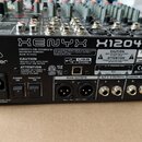 Zadní strana míchacího pultu značky behringer s nápisem XENYX X1204USB. Zařízení má černý kovový povrch s bílými popisky a obsahuje různé konektory,…