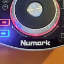 Detailní pohled na část DJ kontroléru značky Numark. Je vidět otočný jog s červeným podsvícením, tlačítka CUE a Play/Pause s modrým podsvícením a ovl…