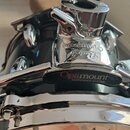 Detailní pohled na horní obruč a mechanismus uchycení černého bubnu. Na chromovaném kovovém kování je vyryt nápis Pearl a pod ním štítek s textem Opt…