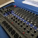 Šedý mixážní pult s modrými prvky, umístěný v otevřeném kufru s modrou výstýlkou. Na ovládacím panelu jsou vidět zahlreiche potenciometry, posuvníky…