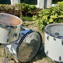 Sada tří bílých bubnů v outdoorovém prostředí. Na levý buben je položen hnědý dřevěný disk, prostřední basový buben má na blaně logo znázorňující kor…