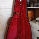 Otevřený hnědý tvrdý kufýr s červeným plyšovým vnitřkem. Na černém upevňovacím pásku je zlatý nápis Gibson. Kufýr vykazuje mírné známky opotřebení na…