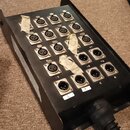 Černý kovový rozvodč (snake box) s řadou female XLR konektorů. Některé konektory jsou přelepeny světlou izolační páskou a u spodních vstupů jsou bílé…