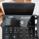 Černý elektronický bicí modul s nápisy BFD a ALESIS DRUMS. Na spodní hraně je uveden model NITRO MAX DRUM MODULE. Zařízení obsahuje displej, ovládací…
