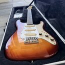 Elektrická kytara s tvarem Stratocaster v hnědo-žlutém sunburst provedení a bílým pickguardem. Na pickguardu je viditelné logo Fender. Nástroj je umí…