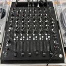 Černý DJ mixér s mnoha ovládacími prvky, potenciometry a posuvníky, umístěný v transportním kufru s hliníkovým rámem. Na panelu jsou viditelné nápisy…