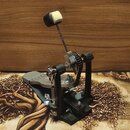 Černý basový pedál s kovovými mechanismy a řetězovým pohonem. Na horní části je umístěn černý beater s bílým plstěným hrotem. Zařízení stojí na černé…