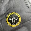 Detailní záběr na černý textilní povrch s kulatou žluto-černou nálepkou. Na nálepce je nápis ROCKBAG by Warwick, proudly made with RokTex a QUALITY B…