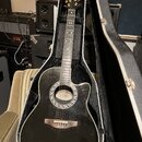 Černá akustická kytara s cutawayem v tvrdém kufru. Kolem otvoru rezonátoru je vidět ornamentální bílá dekorace a nápis "Squier". Hmatník má vložky ve…