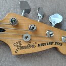 Detail hlavy basové kytary z přírodního dřeva s nápisem Fender® MUSTANG® BASS. Na hlavě jsou čtyři chromované ladicí strojky a čtyři struny vedené př…