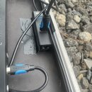 Fotografie zachycuje černý USB hub s připojeným modrým USB konektorem, umístěný v transportním kufru. Na hubu je viditelný nápis "al". V popředí je č…