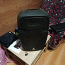 Černý reproduktor s plastovým krytem a mřížkou, umístěný na světlém dřevěném stolku. Ve spodní části mřížky je viditelné logo s písmenem X a nápisem…
